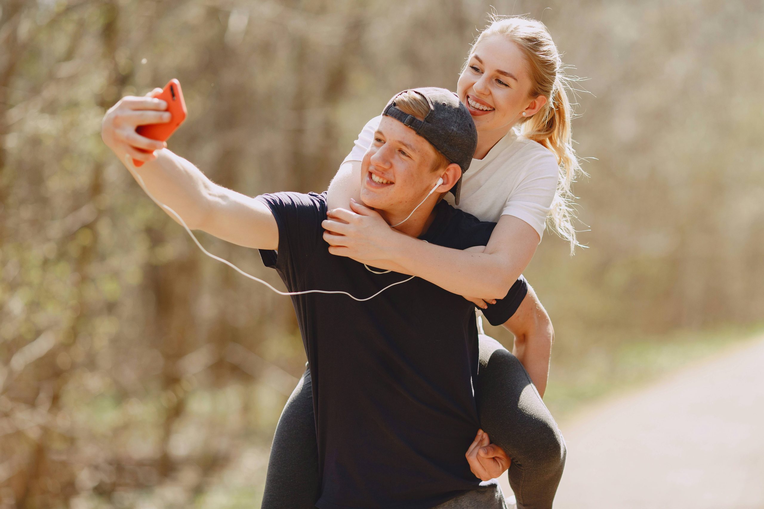 A young man gives a piggyback ride to a young woman as they take a selfie together outdoors, both wearing earphones and casual clothing, capturing one of their playful couple photos.