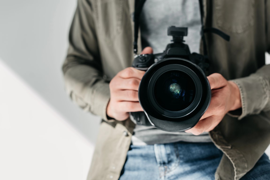 A person stands confidently, holding a professional camera and pointing it towards the viewer, embodying the vibrant energy of Perth photography. Dressed in a casual jacket and jeans, they capture moments that tell stories beyond the lens.